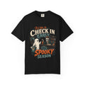 A black short-sleeve crewneck t-shirt featuring a colorful graphic print on the front with the text 'I'd like to CHECK IN EARLY SPOOKY SEASON' and illustrations of a skeleton concierge, a ghost, and a haunted house.