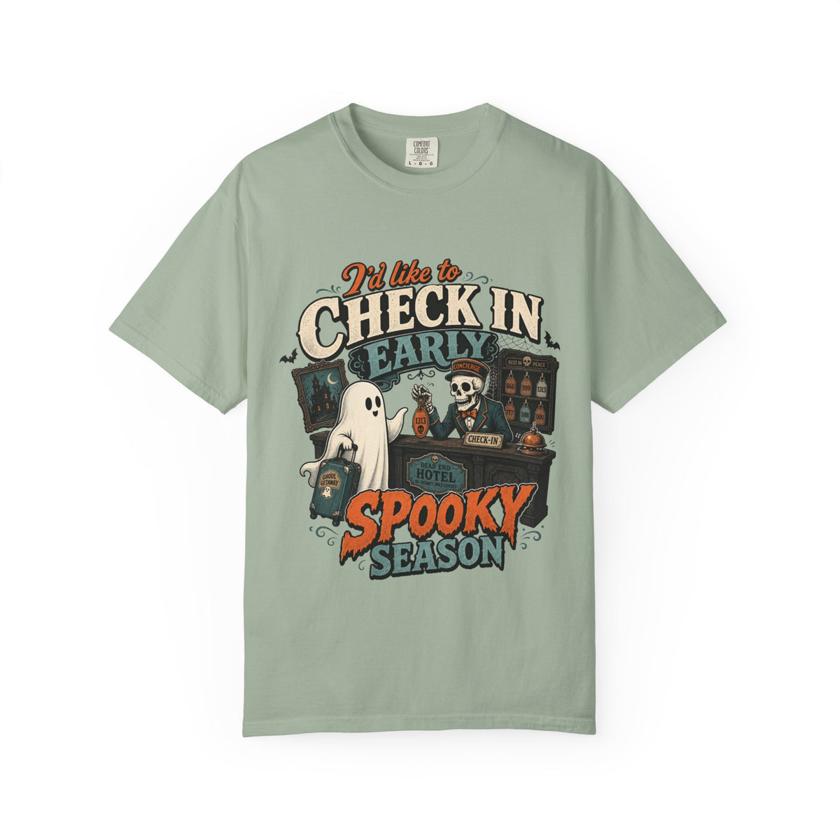A sage green short-sleeve crewneck t-shirt featuring a colorful graphic print on the front with the text 'I'd like to CHECK IN EARLY SPOOKY SEASON' and illustrations of a skeleton concierge, a ghost, and a haunted house.