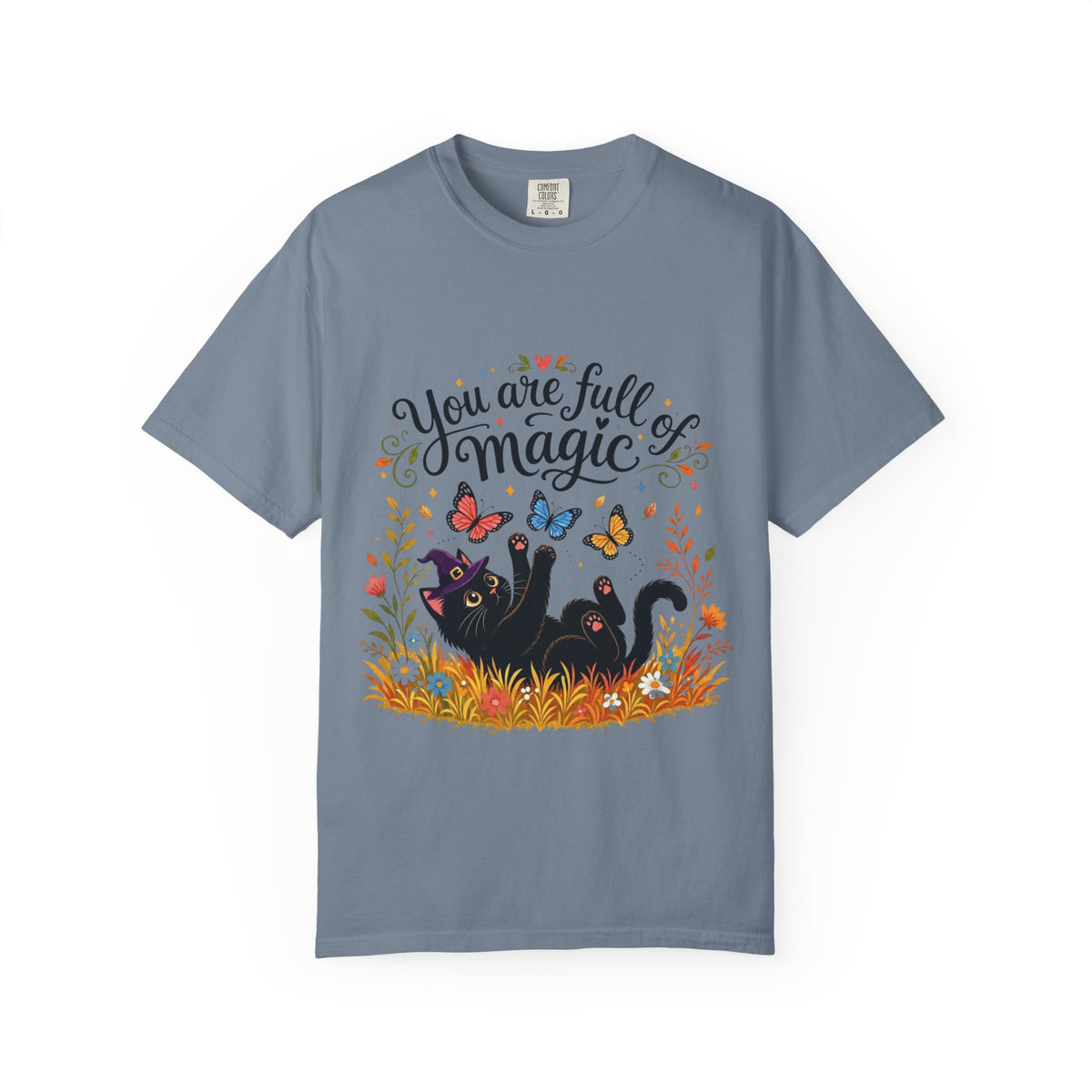 A slate blue short-sleeve crewneck t-shirt featuring a colorful graphic print on the front. The design includes the text 'You are full of magic' in a black script font, above an illustration of a black cat surrounded by colorful butterflies, flowers, and grass.