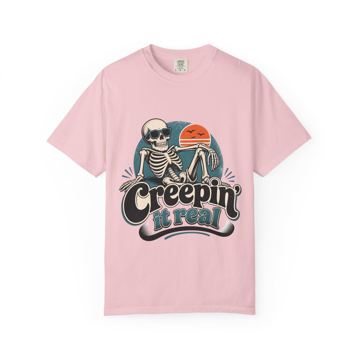 A light pink short-sleeve crewneck t-shirt featuring a retro graphic of a skeleton sitting next to an orange sun, with the text 'Creepin' it real' printed below.