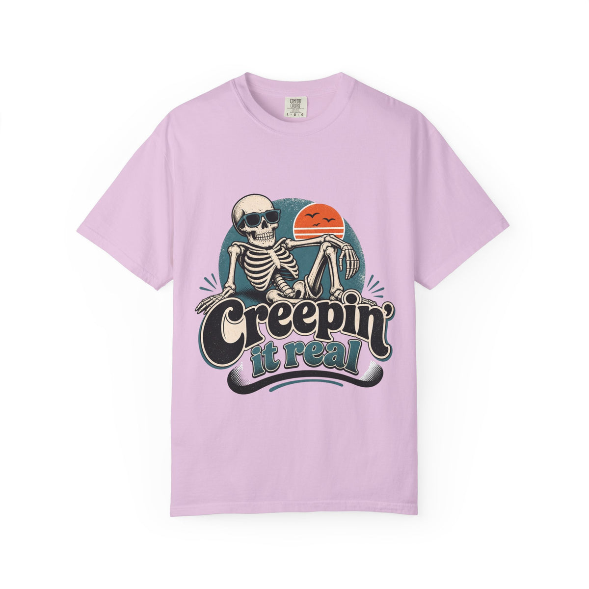 A pink short-sleeve crewneck t-shirt featuring a retro graphic of a skeleton sitting next to an orange sun, with the text 'Creepin' it real' printed below.