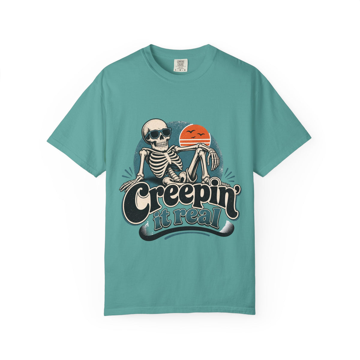 A teal short-sleeve crewneck t-shirt featuring a retro graphic of a skeleton sitting next to an orange sun, with the text 'Creepin' it real' printed below.