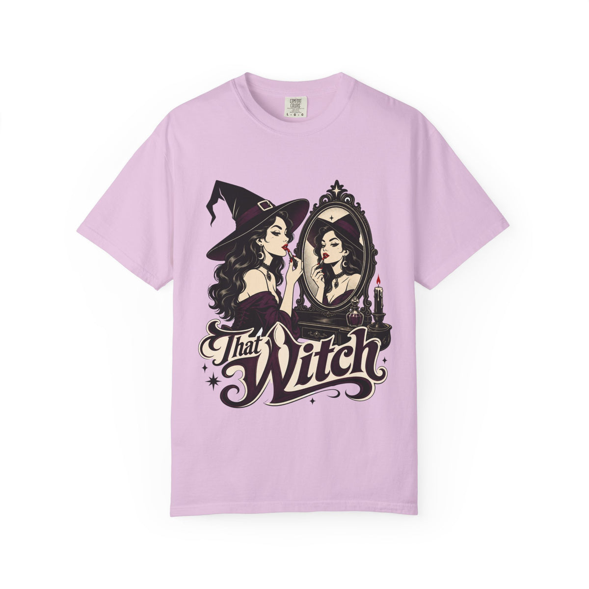 A light pink short-sleeve crewneck t-shirt featuring a graphic print of a witch applying lipstick in front of a mirror, with the text 'That Witch' written below in a stylized font.