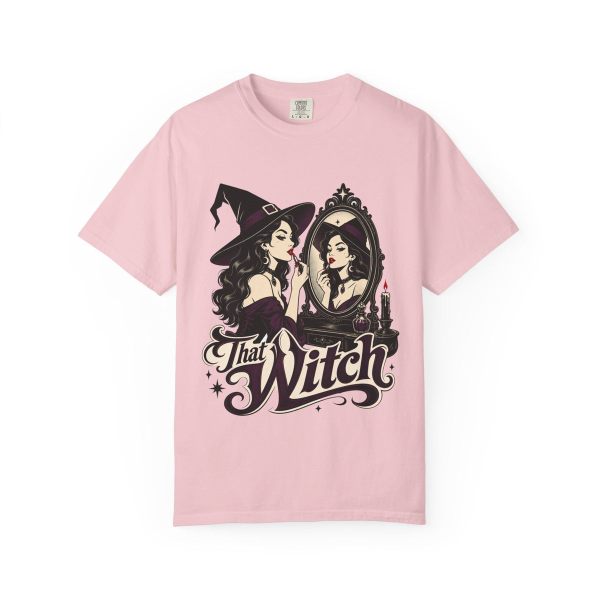 A pink short-sleeve crewneck t-shirt featuring a graphic print of a witch applying lipstick in front of a mirror, with the text 'That Witch' written below in a stylized font.