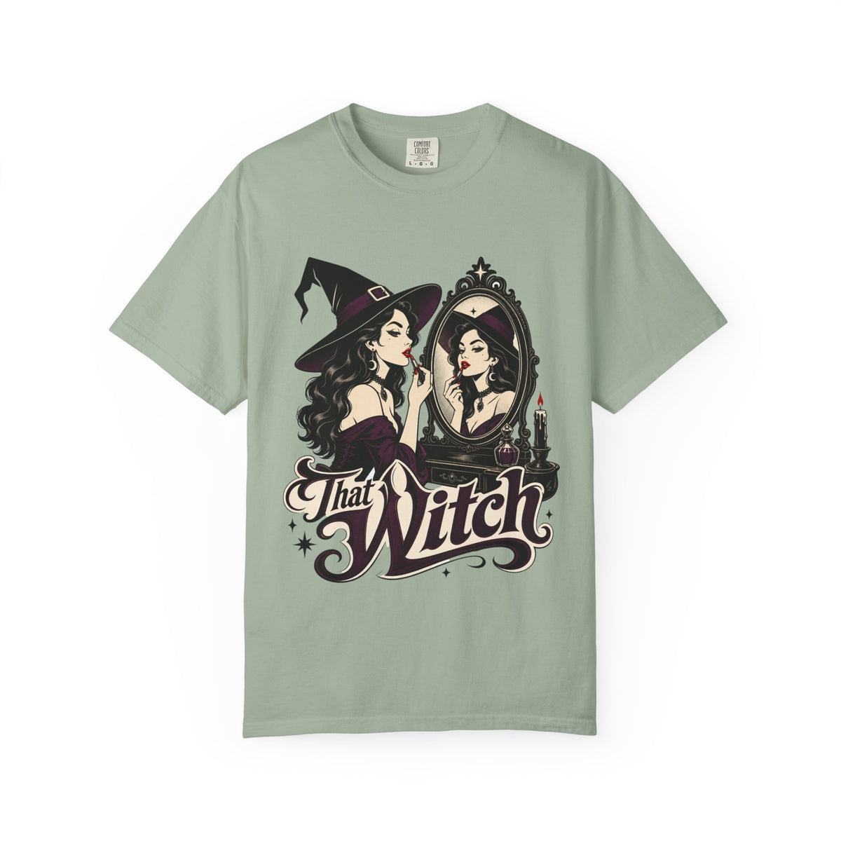 A sage green short-sleeve crewneck t-shirt featuring a graphic print of a witch applying lipstick in front of a mirror, with the text 'That Witch' written below in a stylized font.