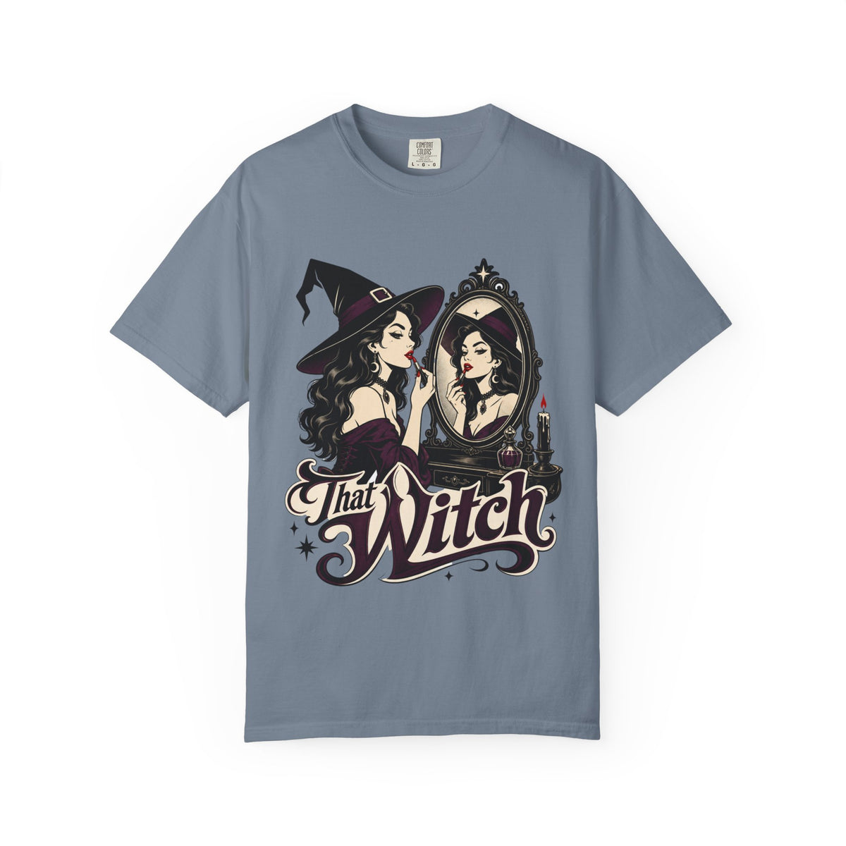 A slate blue short-sleeve crewneck t-shirt featuring a graphic print of a witch applying lipstick in front of a mirror, with the text 'That Witch' written below in a stylized font.