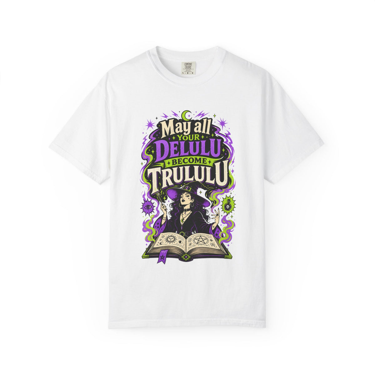 A white short-sleeve crewneck t-shirt featuring a colorful graphic print on the front. The graphic depicts a witch sitting at a table with an open book, surrounded by purple and green decorative elements and the text 'May all your DELULU BECOME Trululu'.