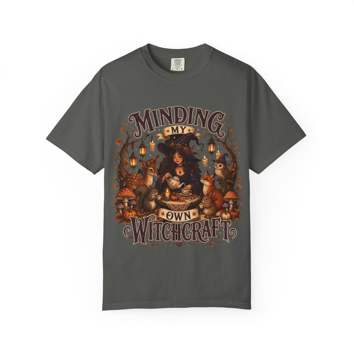 A dark gray short-sleeve crewneck t-shirt featuring a large, detailed graphic that depicts a witch surrounded by autumn elements like foxes, a cat, pumpkins, and lanterns, with the text 'MINDING MY OWN WITCHCRAFT' in a stylized font.