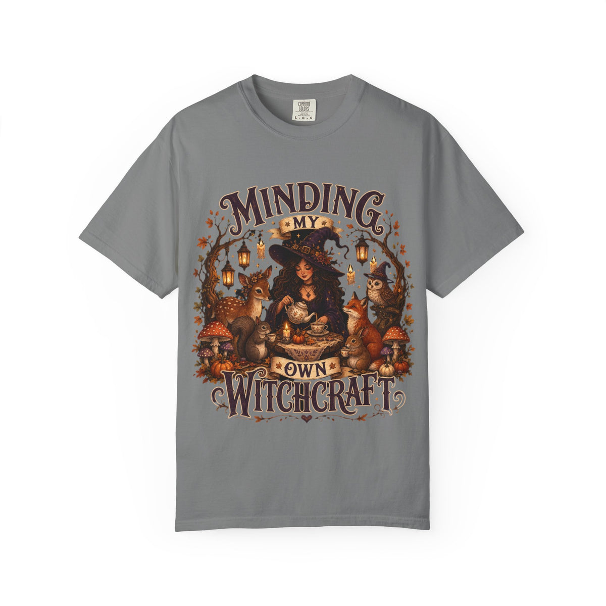 A gray short-sleeve crewneck t-shirt featuring a large, detailed graphic that depicts a witch surrounded by autumn elements like foxes, a cat, pumpkins, and lanterns, with the text 'MINDING MY OWN WITCHCRAFT' in a stylized font.
