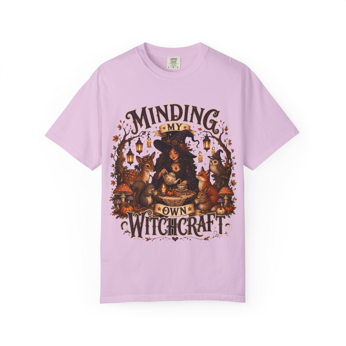 A light purple short-sleeve crewneck t-shirt featuring a large, detailed graphic that depicts a witch surrounded by autumn elements like foxes, a cat, pumpkins, and lanterns, with the text 'MINDING MY OWN WITCHCRAFT' in a stylized font.