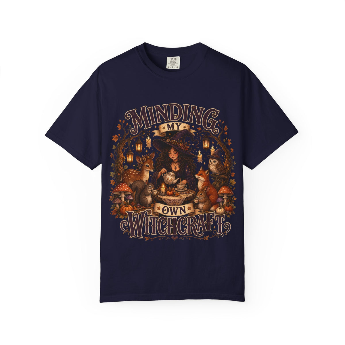 A navy blue short-sleeve crewneck t-shirt featuring a large, detailed graphic that depicts a witch surrounded by autumn elements like foxes, a cat, pumpkins, and lanterns, with the text 'MINDING MY OWN WITCHCRAFT' in a stylized font.