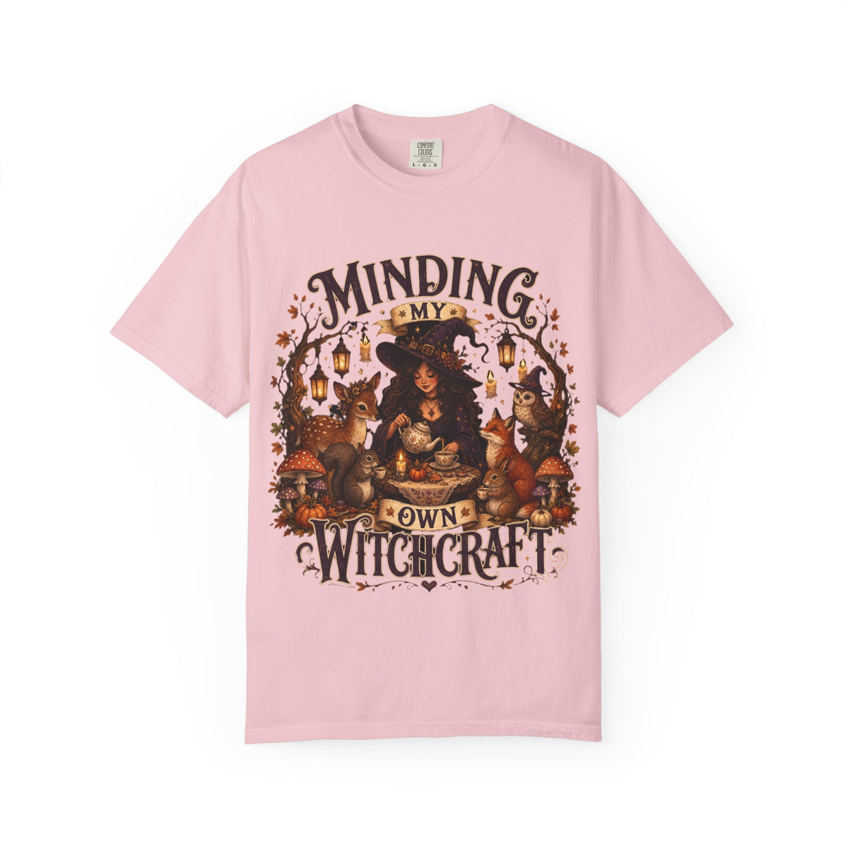 A pink short-sleeve crewneck t-shirt featuring a large, detailed graphic that depicts a witch surrounded by autumn elements like foxes, a cat, pumpkins, and lanterns, with the text 'MINDING MY OWN WITCHCRAFT' in a stylized font.