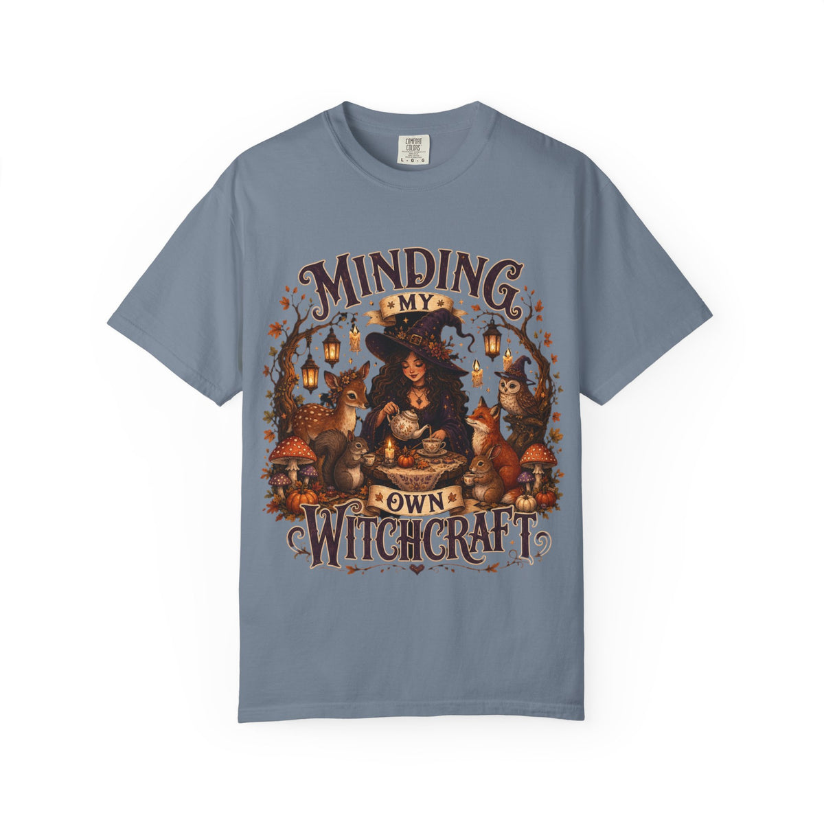 A slate blue short-sleeve crewneck t-shirt featuring a large, detailed graphic that depicts a witch surrounded by autumn elements like foxes, a cat, pumpkins, and lanterns, with the text 'MINDING MY OWN WITCHCRAFT' in a stylized font.