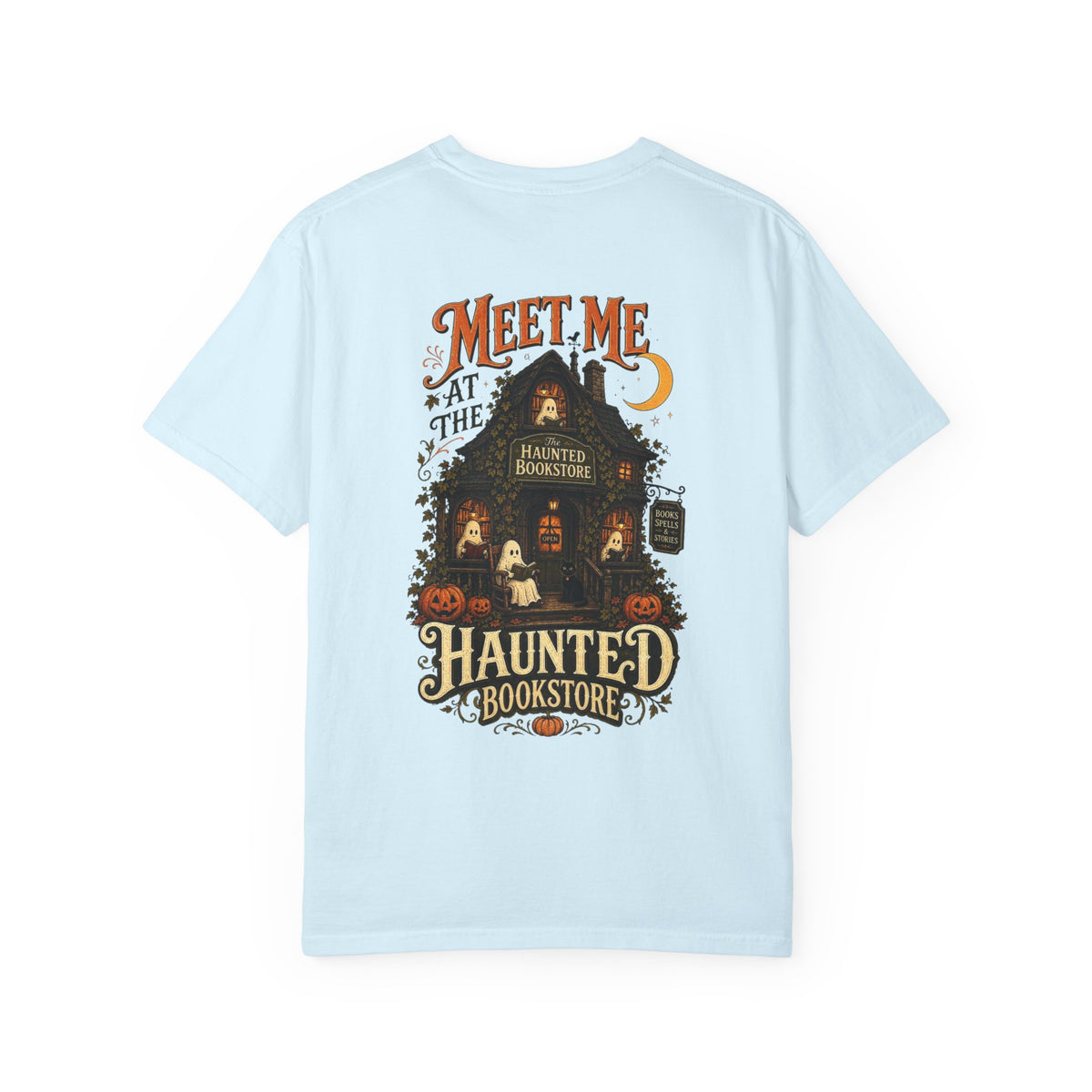A light blue short-sleeve t-shirt with a detailed graphic on the front depicting a haunted bookstore at night, complete with pumpkins, a ghost, and orange text reading 'MEET ME AT THE HAUNTED BOOKSTORE'.