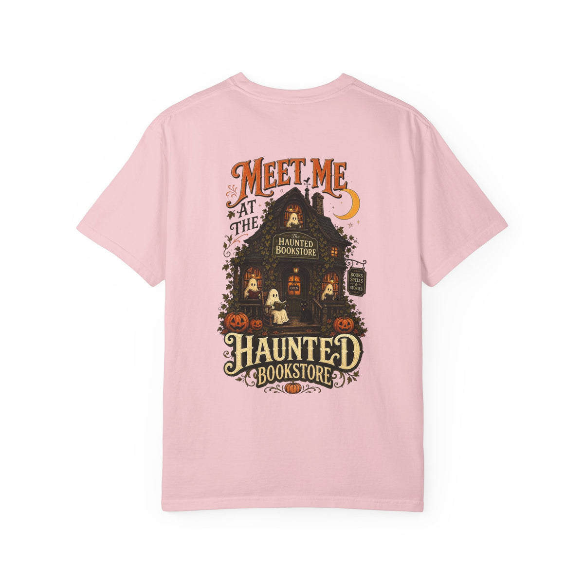 A pink short-sleeve t-shirt with a detailed graphic on the front depicting a haunted bookstore at night, complete with pumpkins, a ghost, and orange text reading 'MEET ME AT THE HAUNTED BOOKSTORE'.