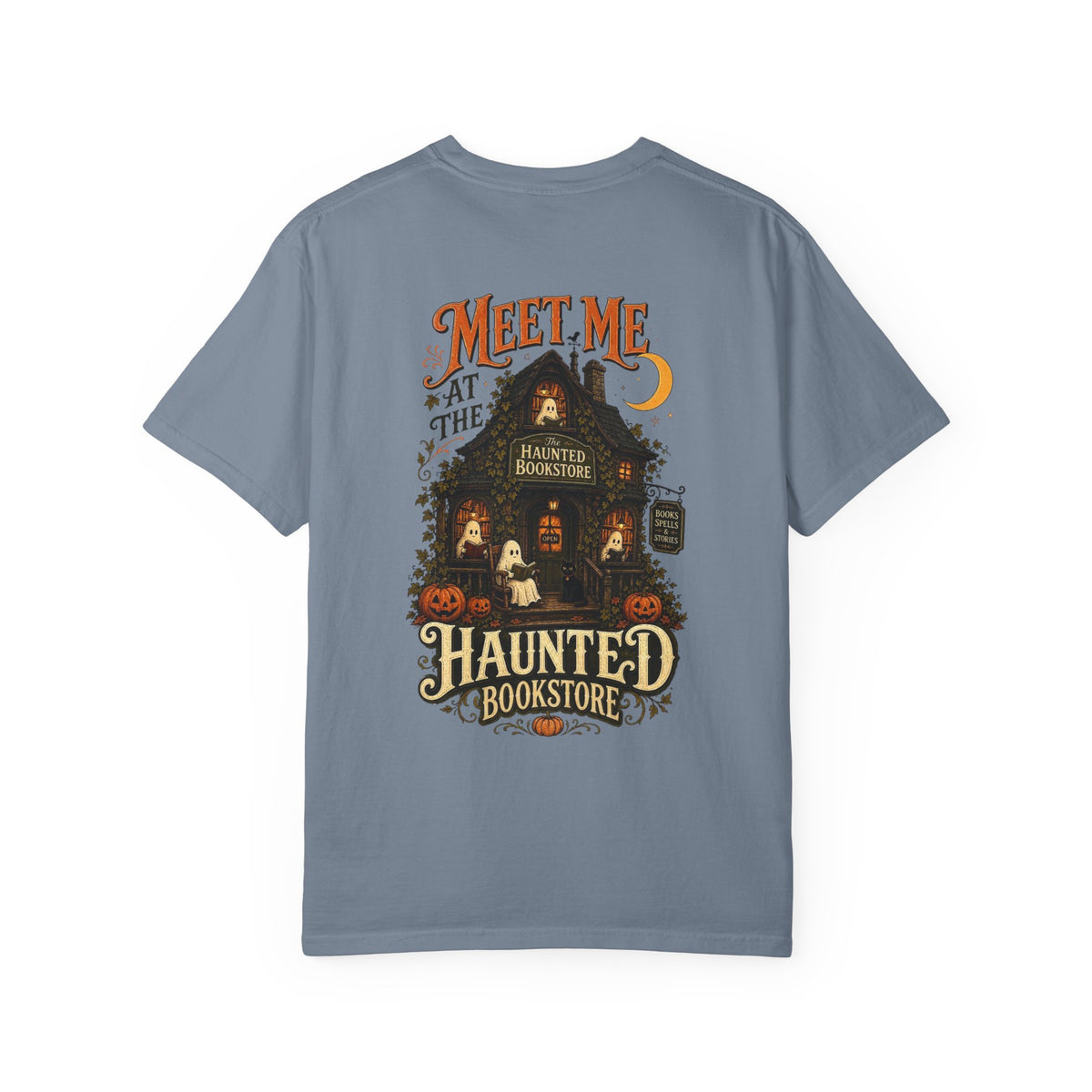 A slate blue short-sleeve t-shirt with a detailed graphic on the front depicting a haunted bookstore at night, complete with pumpkins, a ghost, and orange text reading 'MEET ME AT THE HAUNTED BOOKSTORE'.