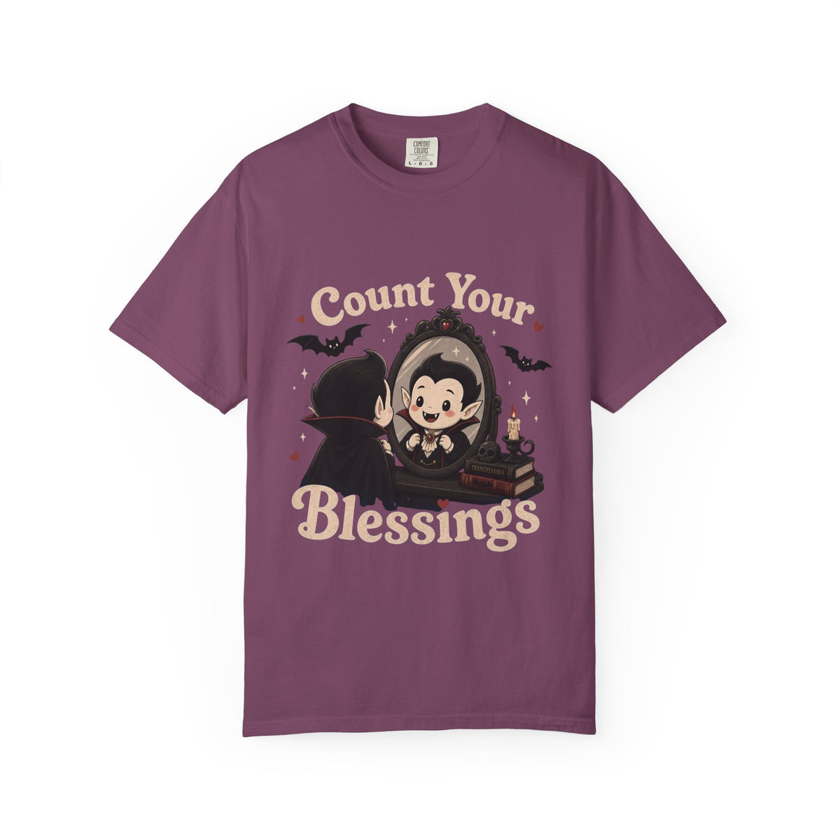 A dark purple short-sleeve crewneck t-shirt featuring a cute cartoon graphic of a vampire smiling at mirror, with the text 'Count Your Blessings' in a peach-colored font.