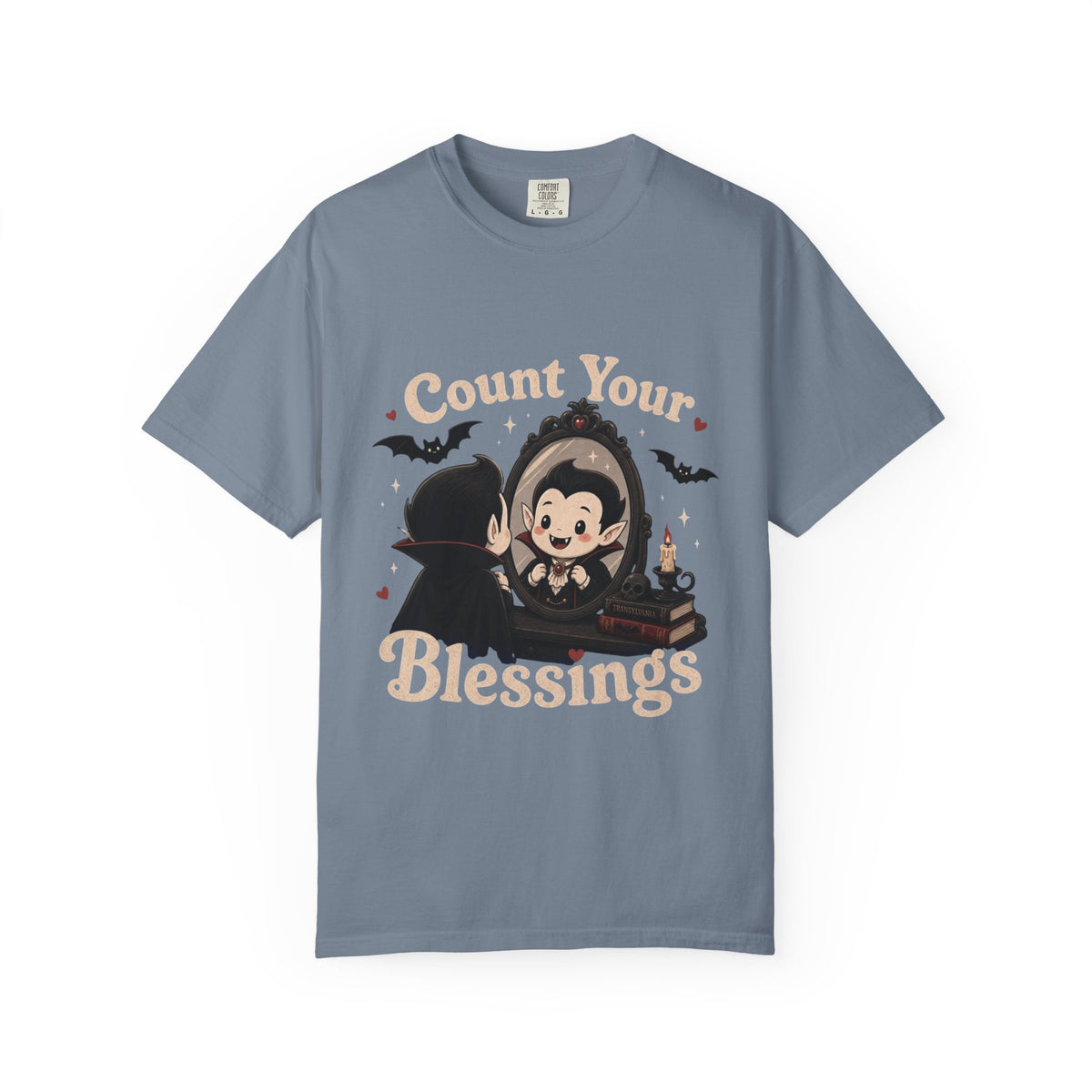 A slate blue short-sleeve crewneck t-shirt featuring a cute cartoon graphic of a vampire smiling at mirror, with the text 'Count Your Blessings' in a peach-colored font.